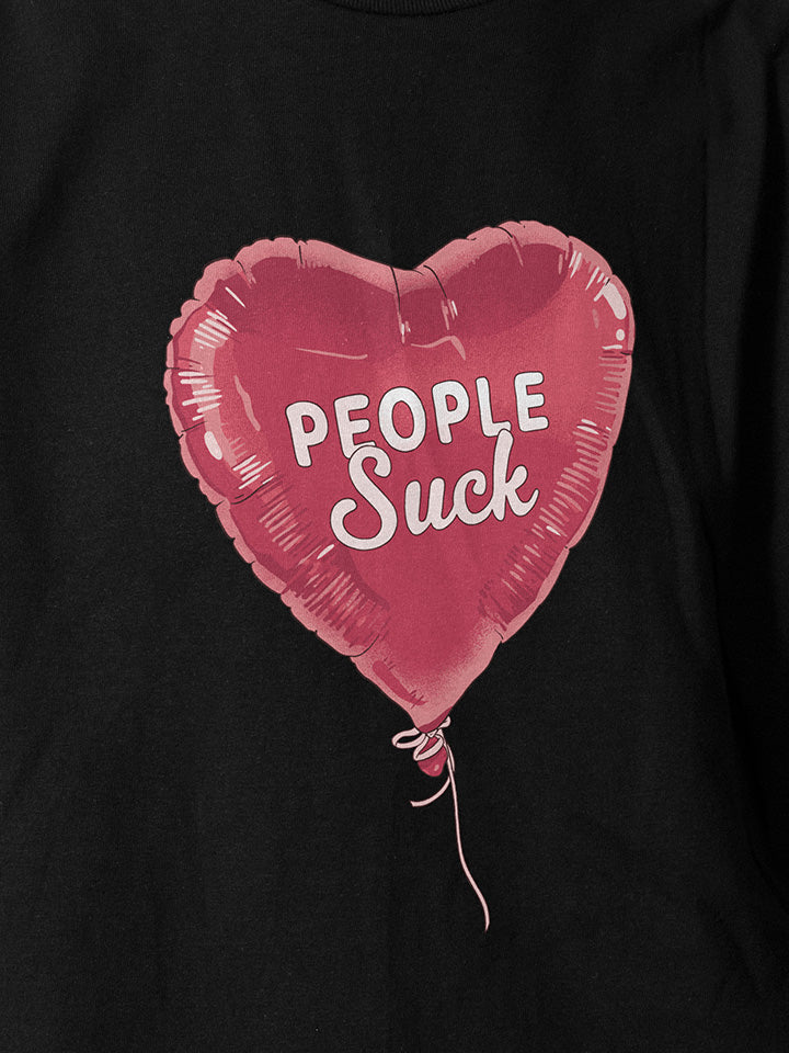 A close-up of a black printed t-shirt by useless treasures. Print of an illustrated heart shapes red balloon with the text people suck.