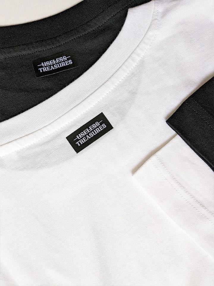 Close-up on two t-shirts showing the high-quality fabric of t-shirts and woven logo label by Berlin-based artist useless treasures available in two colors, black and white. 